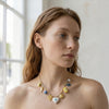 Eda Jewels Dubai gold plated charm necklace featuring eclectic dangling pendants including hearts, a shell, a fish, and evil eye beads, worn by a model near a window.