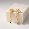 Eda Jewels Dubai gold plated abstract molten earrings with mixed metal fringe drop chains featuring a gold heart padlock, a silver droplet charm, and a black bead accent, displayed on a cream stone block