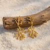 Eda Jewels Dubai gold plated textured sea coral branch drop earrings dotted with tiny white pearl accents, displayed on white sand leaning against a piece of driftwood.
