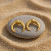 Eda Jewels Dubai gold plated crescent horn stud earrings featuring outer beaded detailing and a central textured dome accent with a single crystal diamond center, displayed on a smooth grey stone in the sand